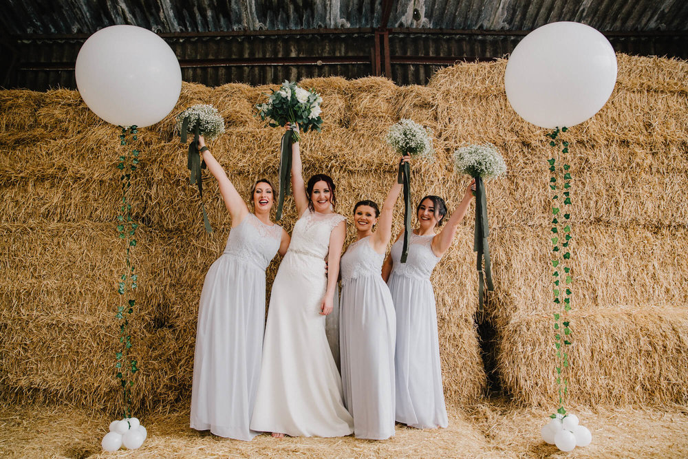 bride and bridal party hold bouquets in the air on top of hay bale backdrop for posed group photograph