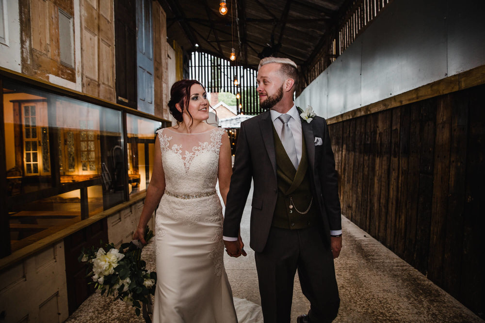 newly wedded couple walking toward camera for portrait flash photograph while holding bouquet against barn door