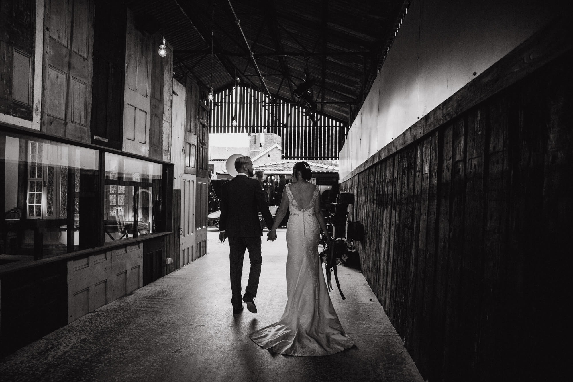 black and white photograph of bride and groom holding hands walking away from camera towards barn entrance hallway