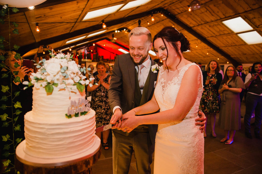 newly married couple cutting into wedding cake surrounded by family and friends