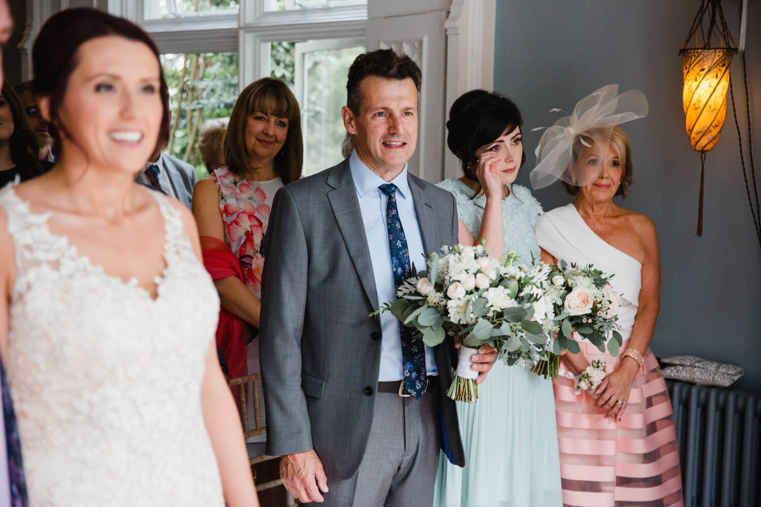 bridesmaid at top of aisle wiping away a tear while father holds bouquet during service