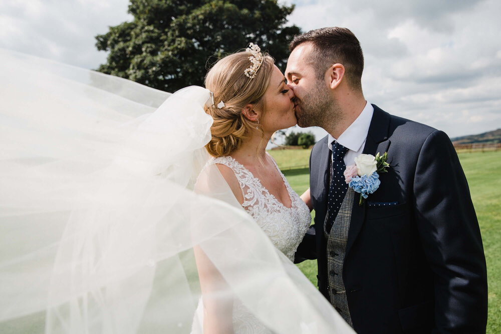 newlyweds kiss under tree as wedding veil blows into foreground