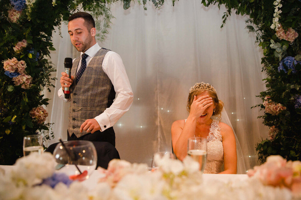 bride with head in hands as groom delivers his speech to wedding party