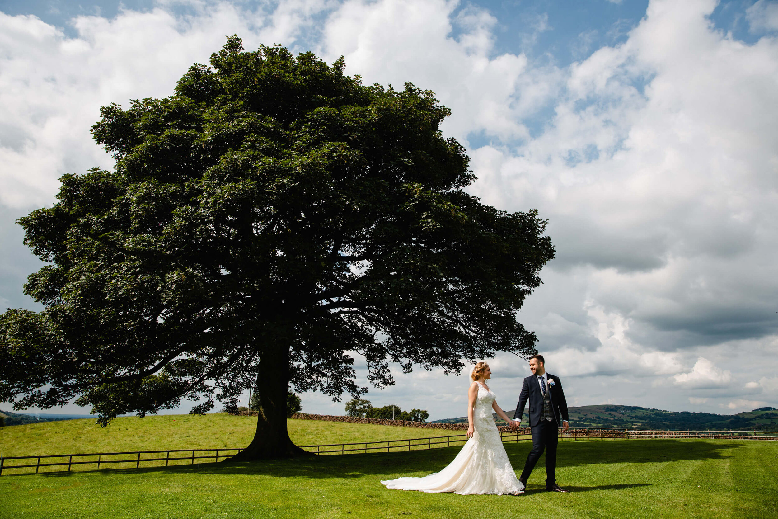 picturesque portrait of newly married couple holding hands on lawn of Heaton House Farm under oak tree