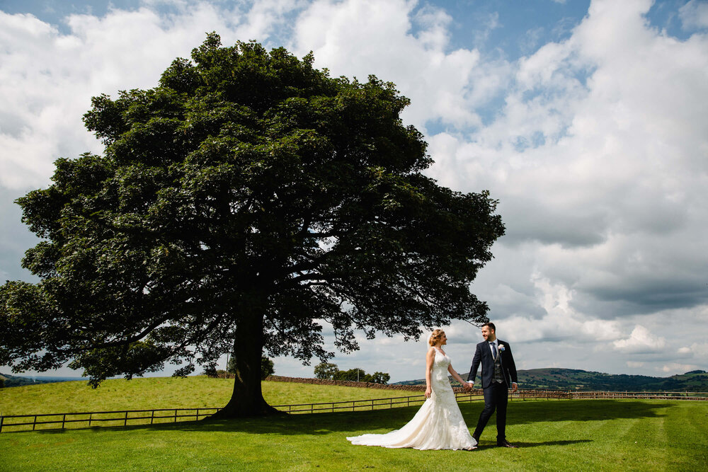 picturesque portrait of newly married couple holding hands on lawn of Heaton House Farm under oak tree