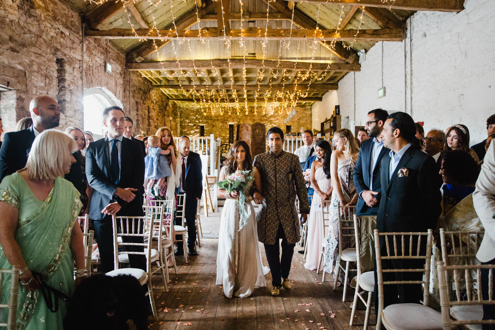 bride walks down barn aisle looking on at groom