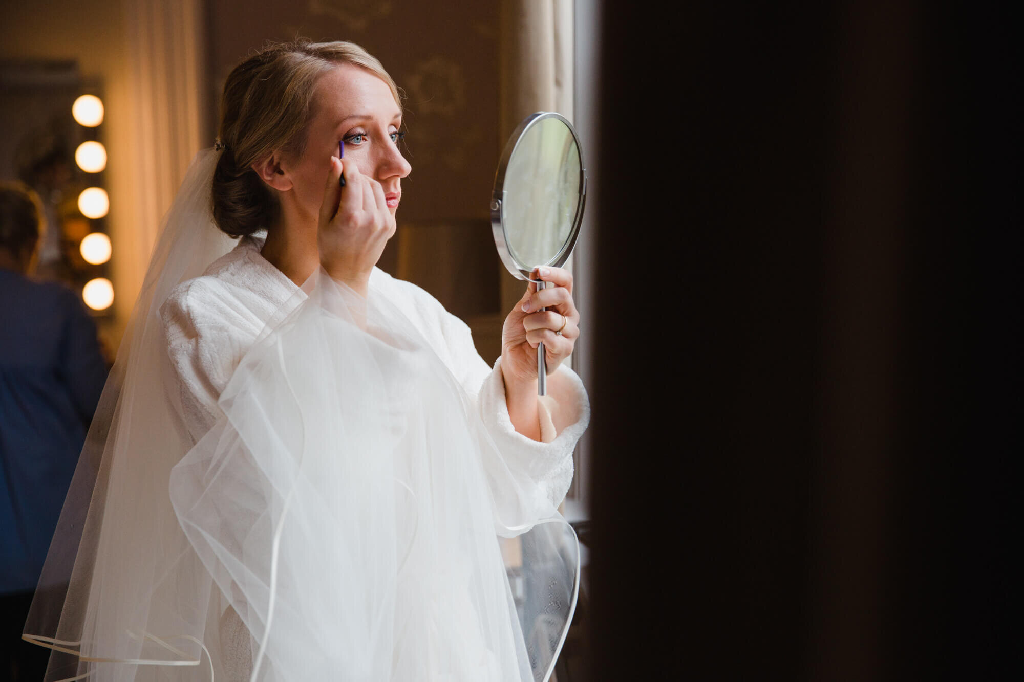 Bride dressed in wedding veil applying mascara in bridal suite mirror 
