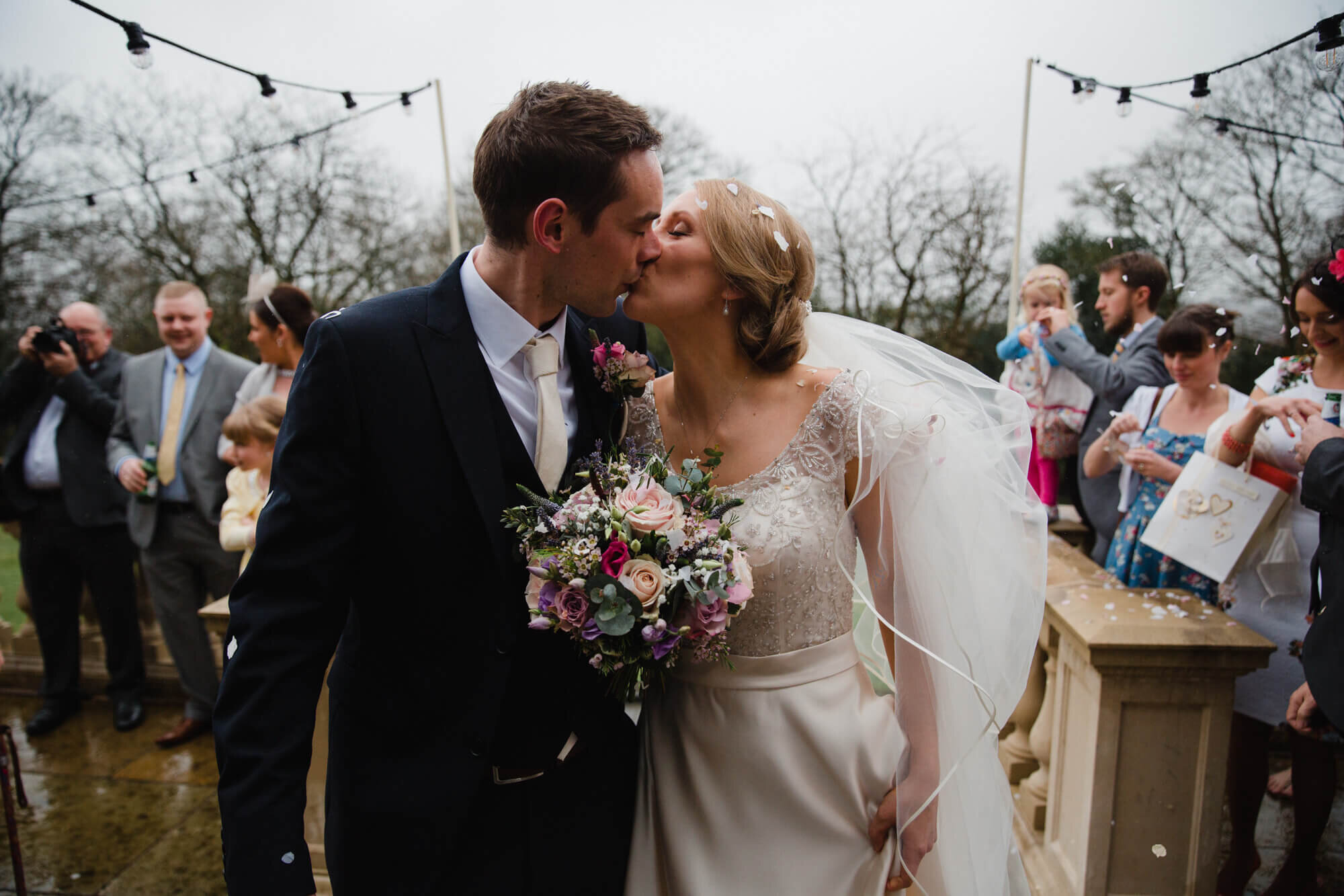 close up first kiss of bride and groom on steps surrounded by friends and family