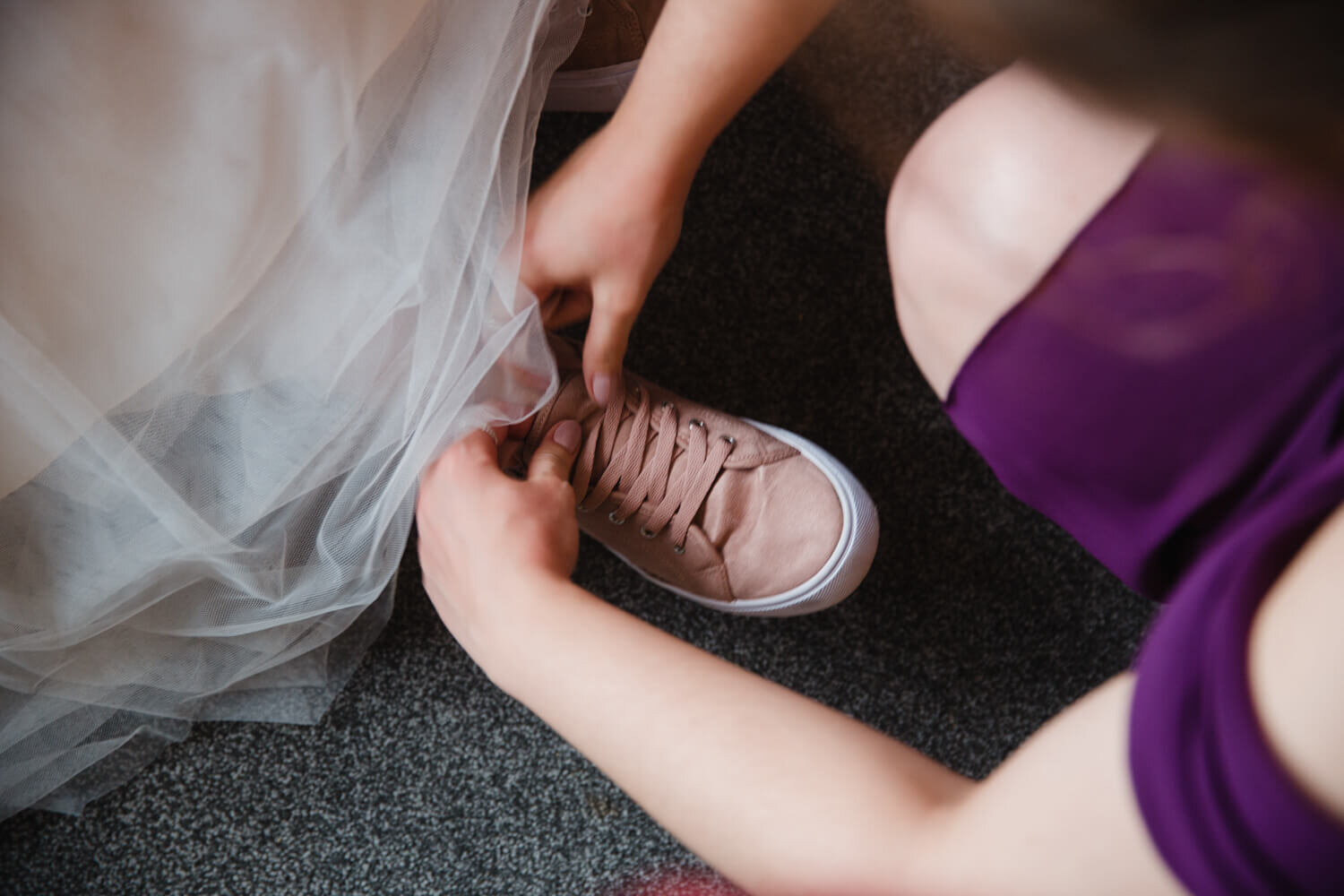 bride wearing pink converse shoes being fastened by bridesmaid