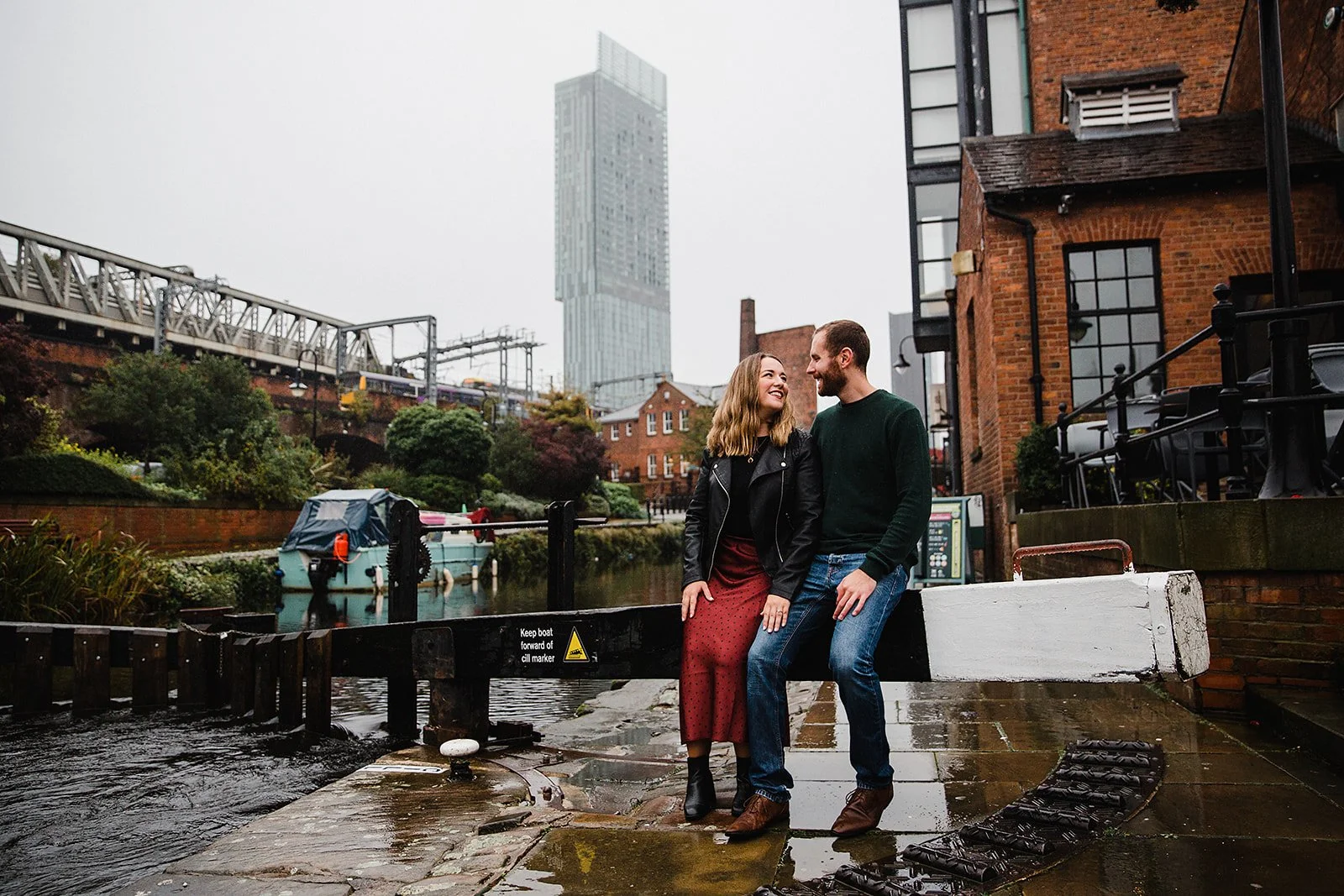 Couple sitting together on a canal lock gate in Castlefield with the Manchester skyline behind them during a relaxed Deansgate Castlefield couples photography session.