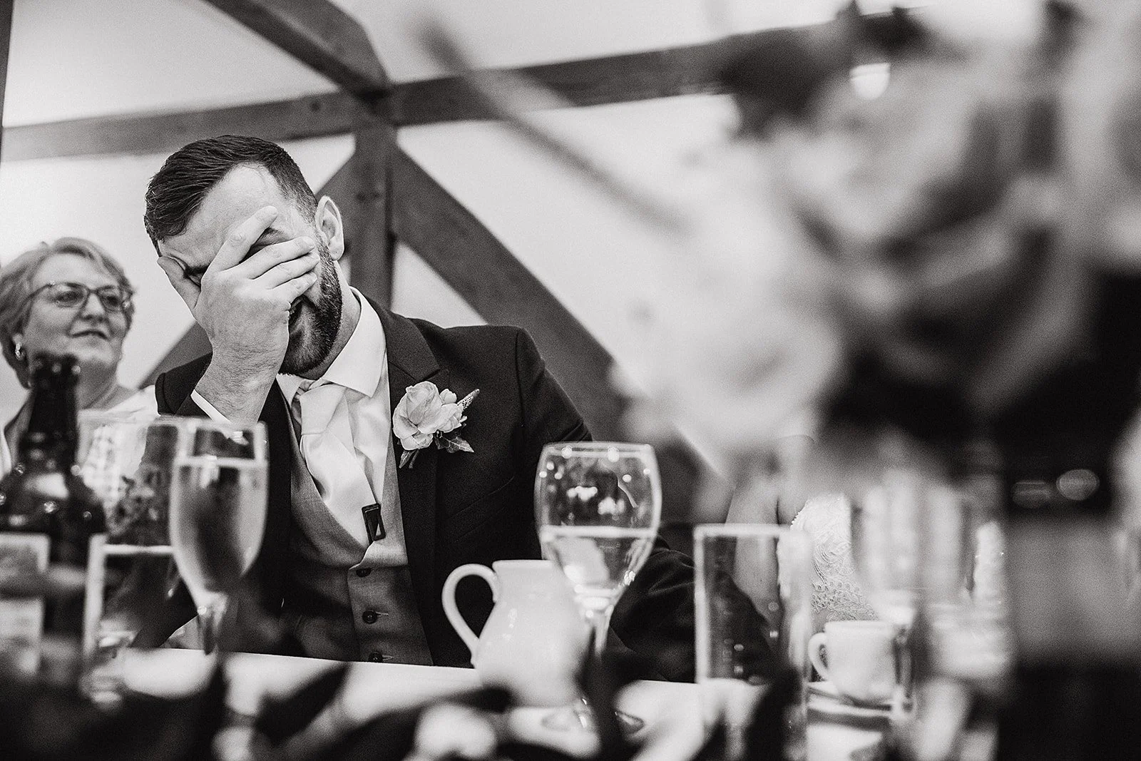 A black and white photo from the top table at a wedding dinner. A groom in a suit and waistcoat sits with his left hand covering his face during the speeches beneath the angled timber beams of the barn.