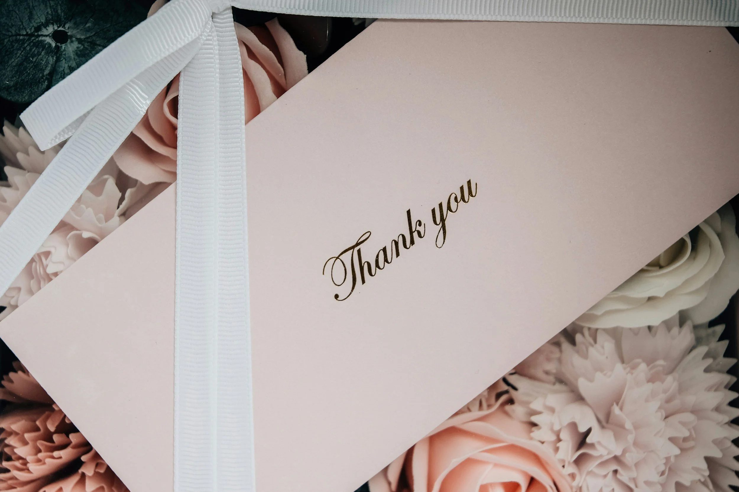A light pink thank you card placed on top of a bouquet of pink and white flowers, tied with a white ribbon.
