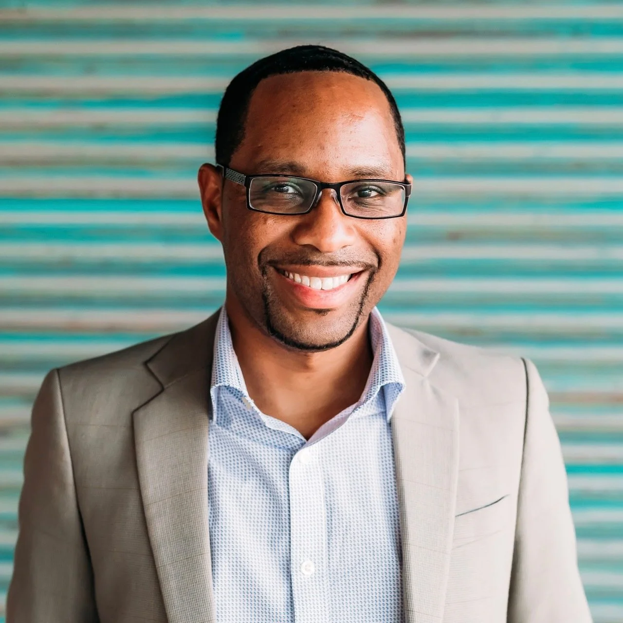 Reuben wearing glasses and a beige suit jacket, smiling in front of a turquoise striped background.