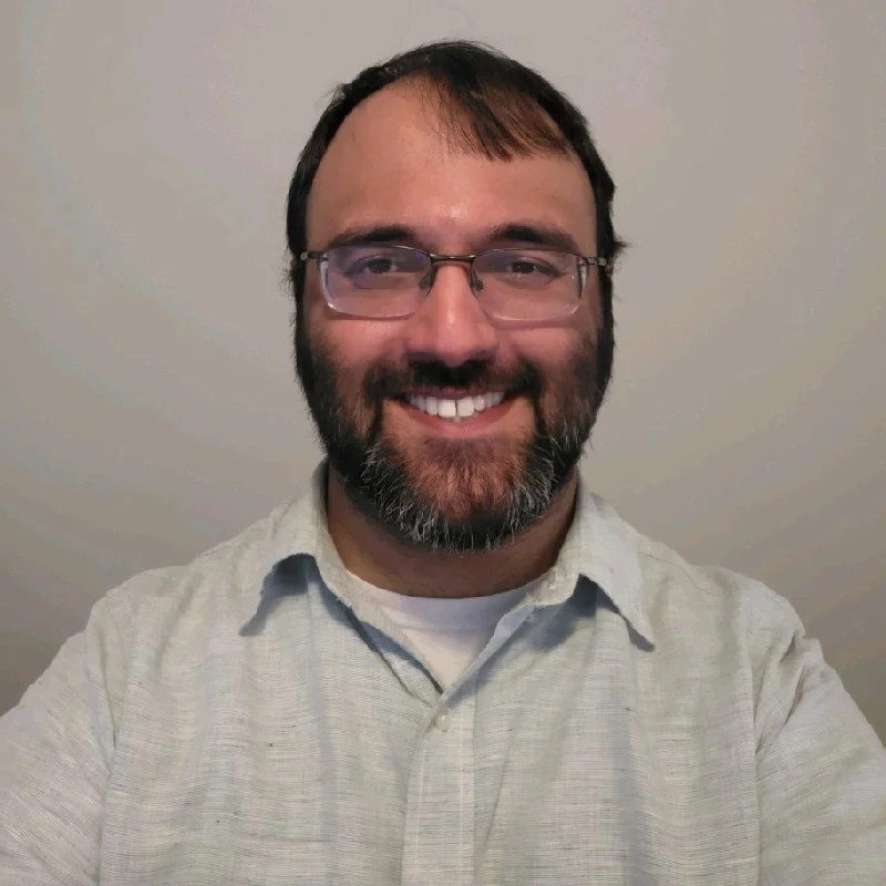 Mo smiling with glasses and beard wearing a light-colored shirt, set against a neutral background.