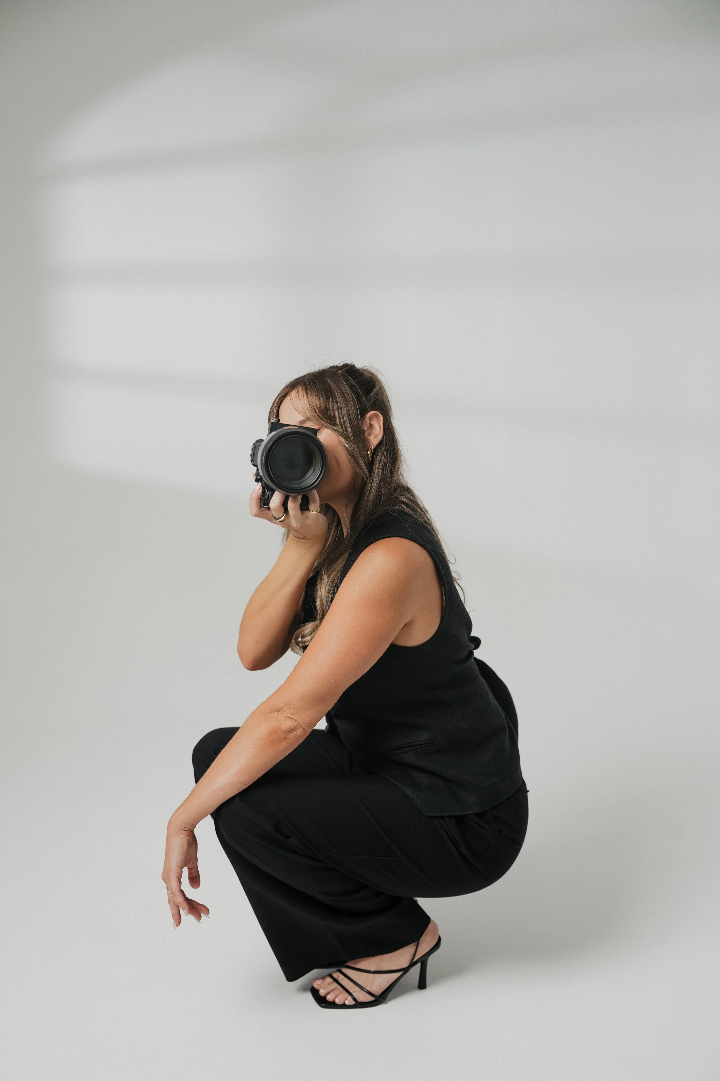 Woman dressed in black, squatting while holding a camera in front of her face, against a plain, light gray background.