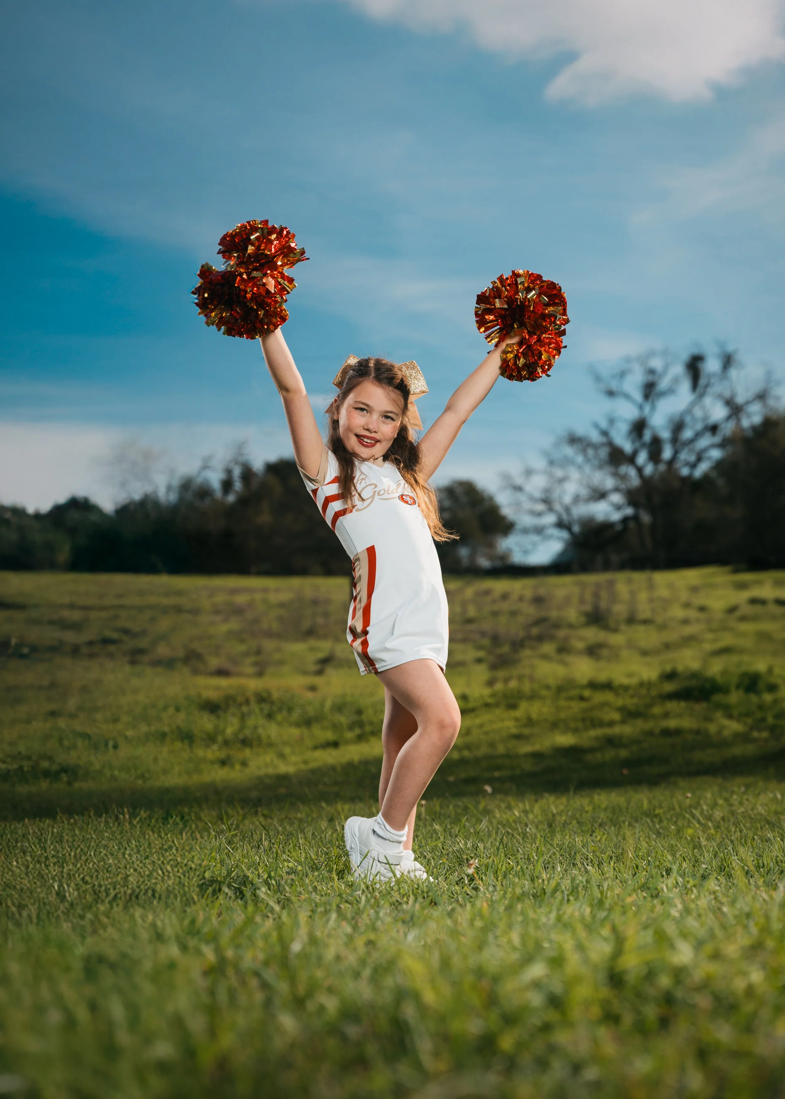 A young girl dressed in a cheerleading outfit standing on grass with arms raised holding red and gold pom-poms, outdoors with a blue sky and trees in the background.