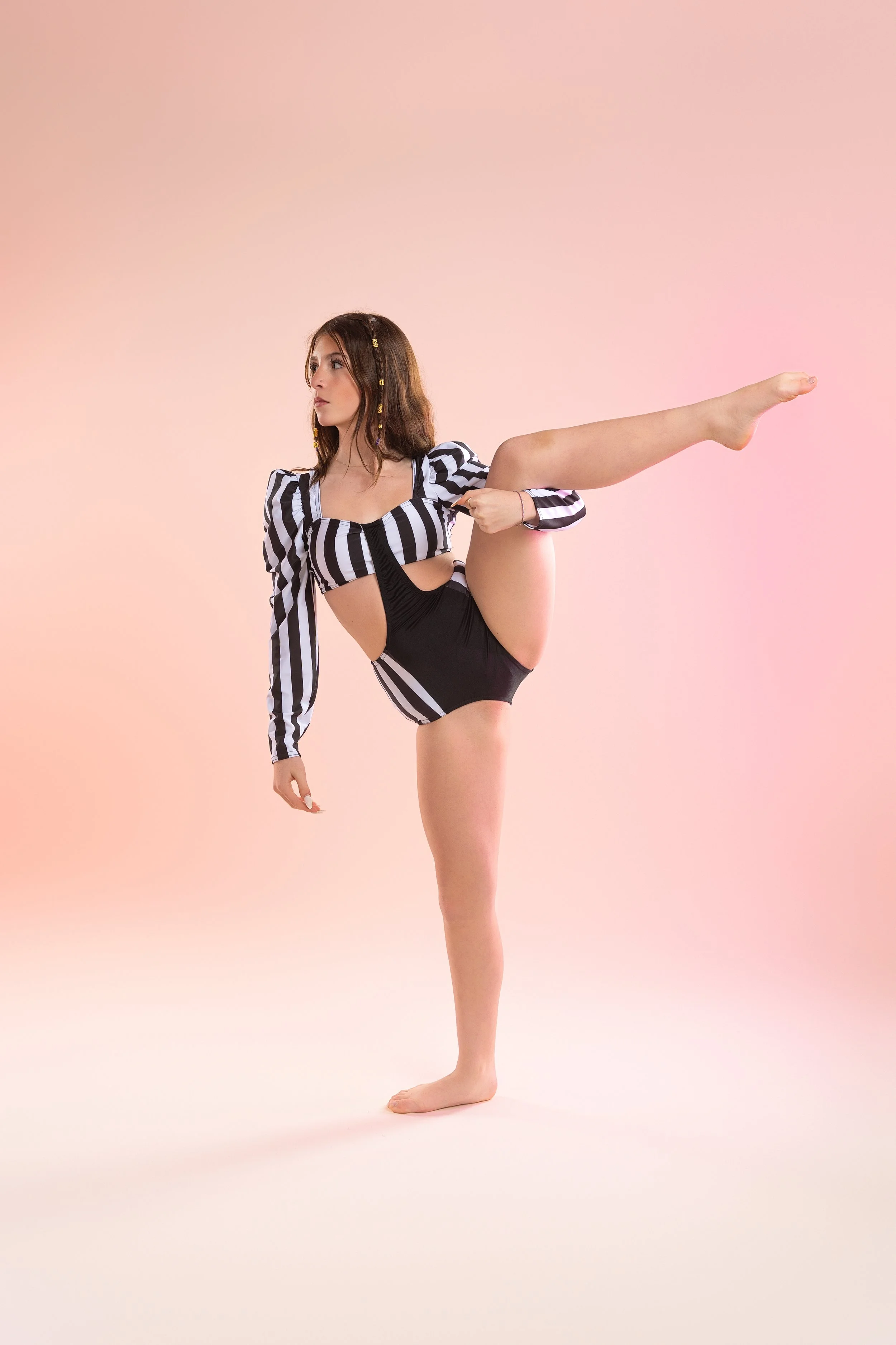 A young woman in a black and white striped dance costume is striking a ballet pose, standing on one leg with the other extended to the side, against a pink gradient background.