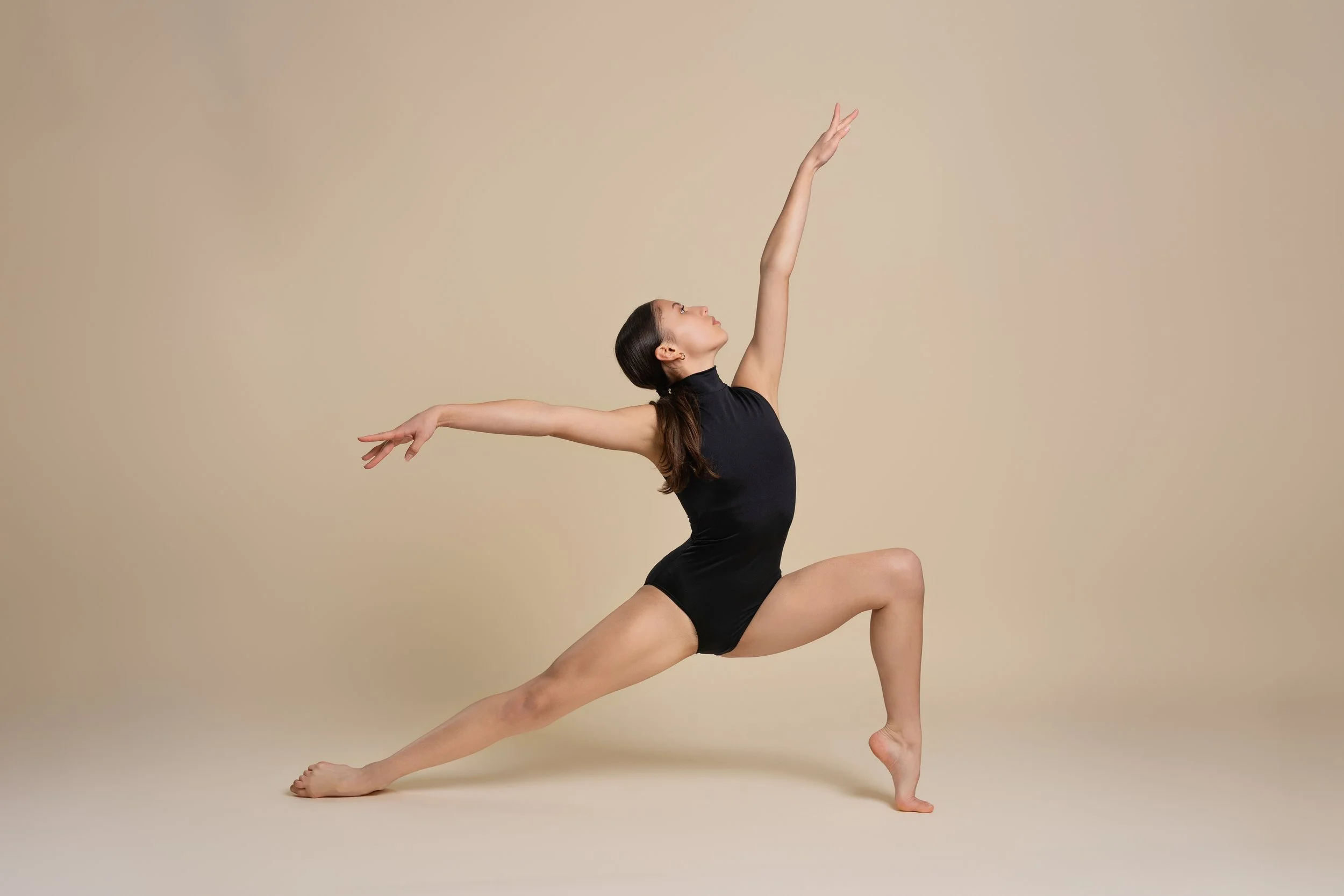 A woman in a black sleeveless leotard performing a yoga pose with one leg bent, one arm reaching forward and the other arm extended upward against a plain beige background.