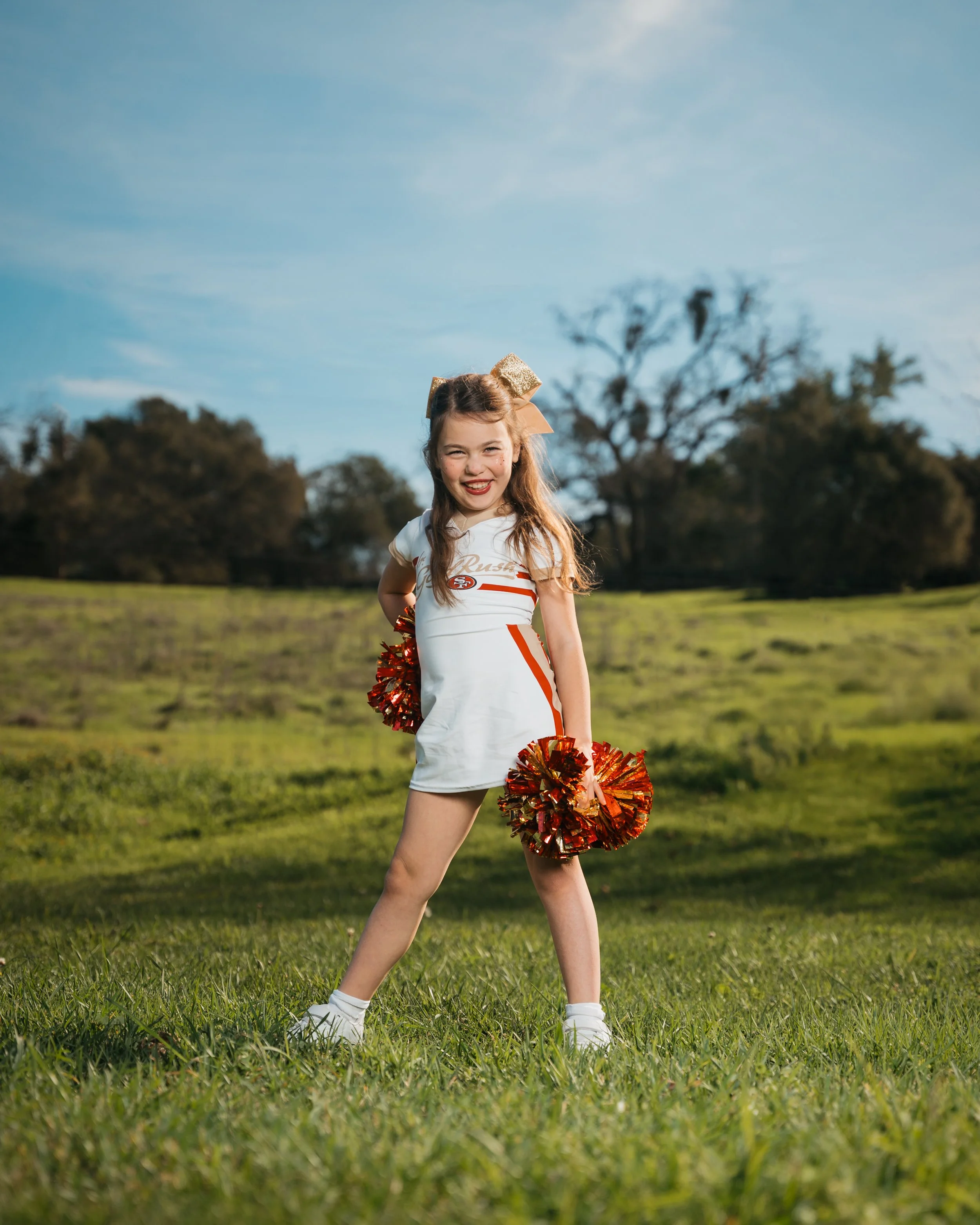 Young girl cheerleader standing outdoors on grass field, holding red and gold pom-poms, smiling, wearing a white cheerleading uniform with a gold bow in her hair.