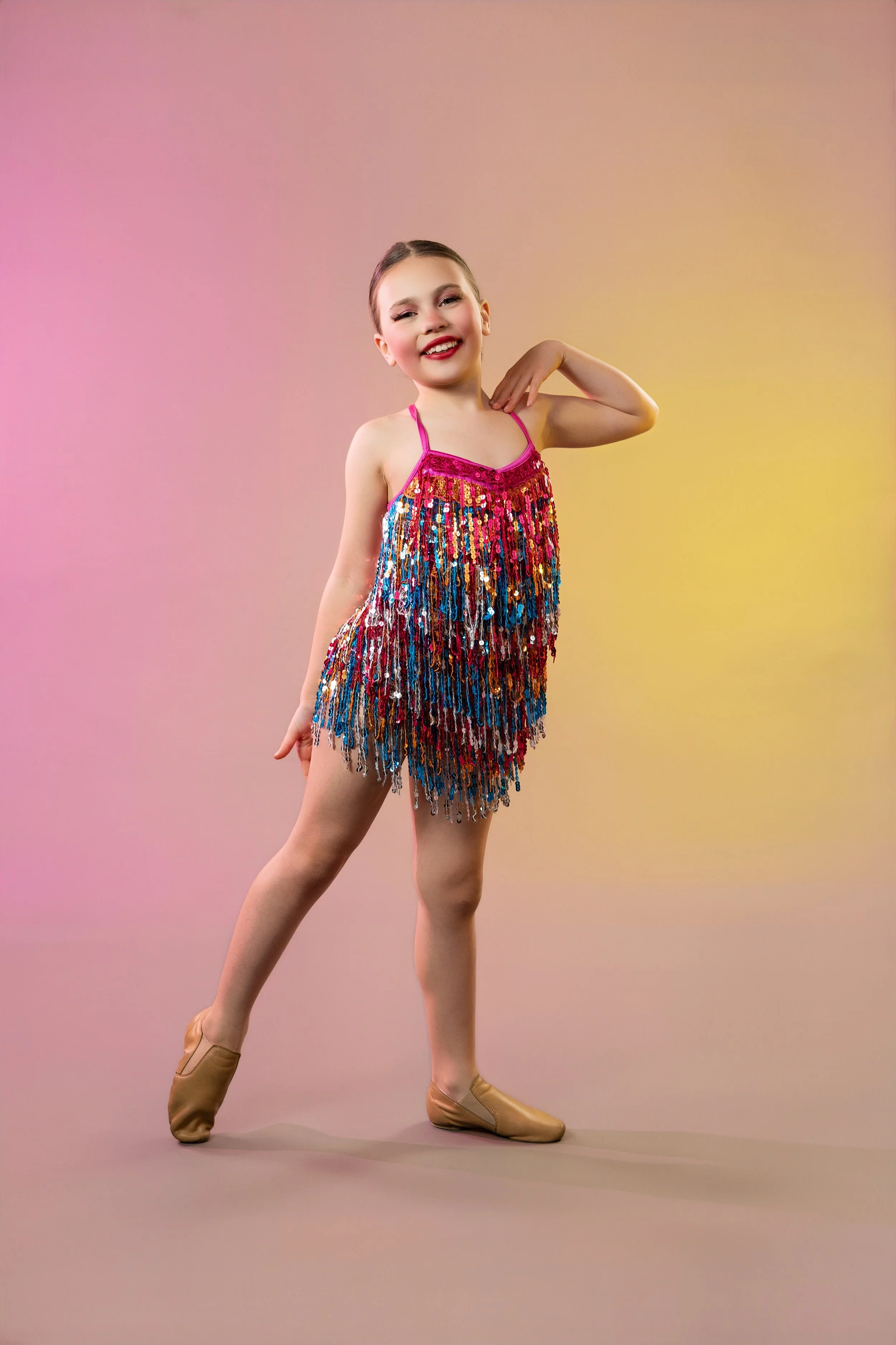 Young girl in a sparkly, multicolored dance costume with gold dance shoes, smiling and posing on a pastel gradient background.