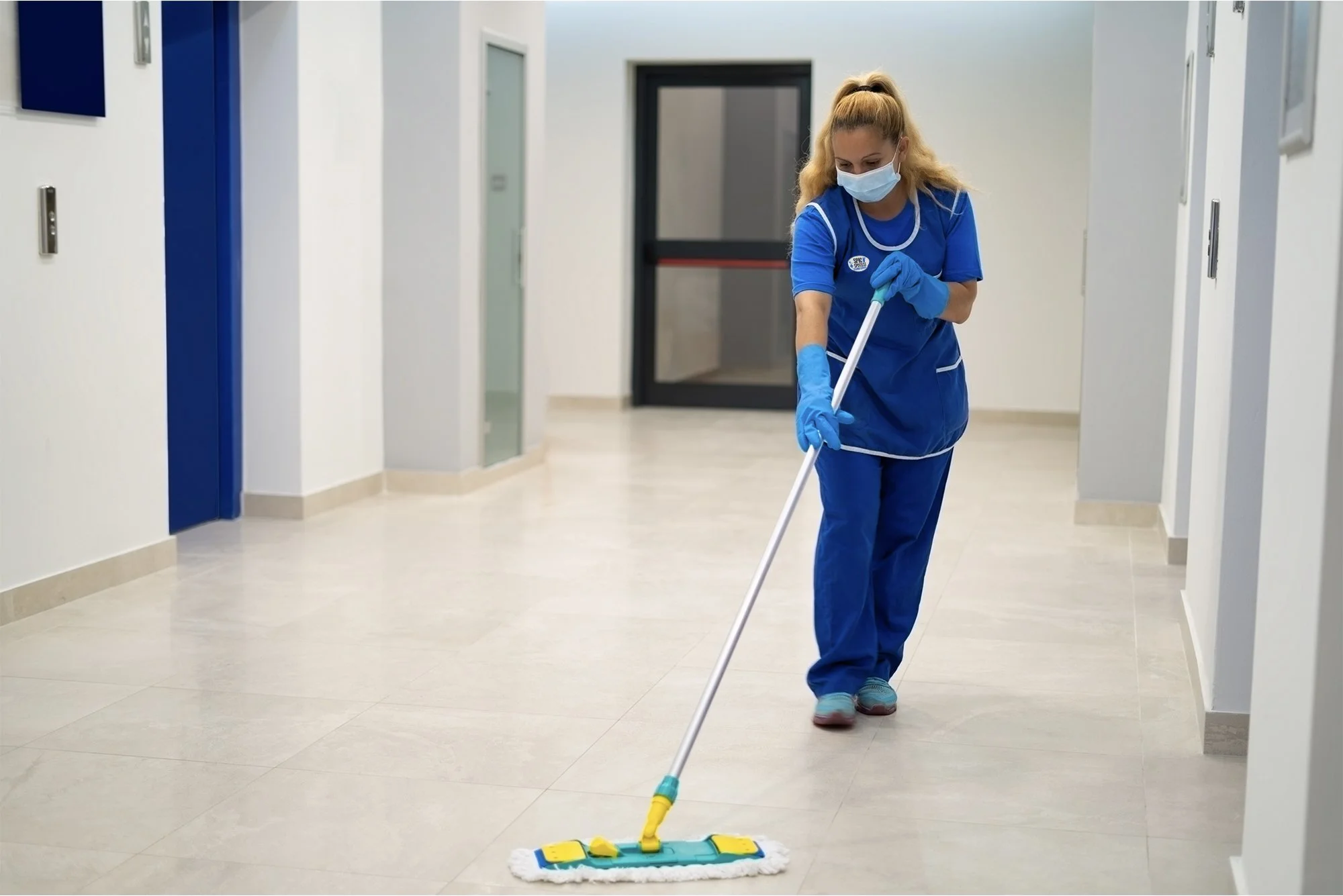 A woman wearing a face mask, gloves, and blue scrubs mops the floor with a cleaning mop in a corridor of a building, possibly a hospital or clinic.
