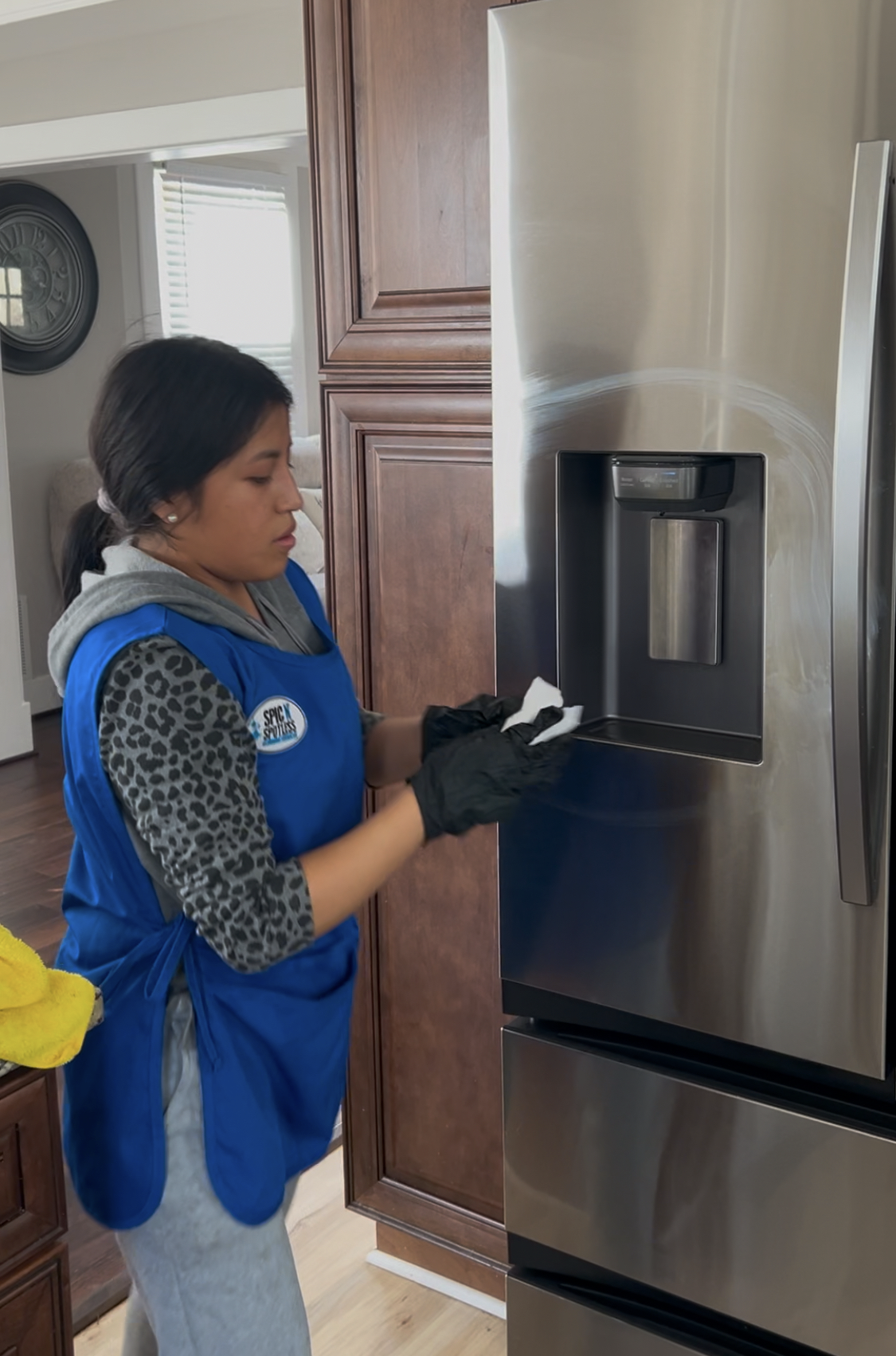 A woman wearing a blue apron and black gloves cleaning the inside of a stainless steel refrigerator with a cloth in a kitchen.