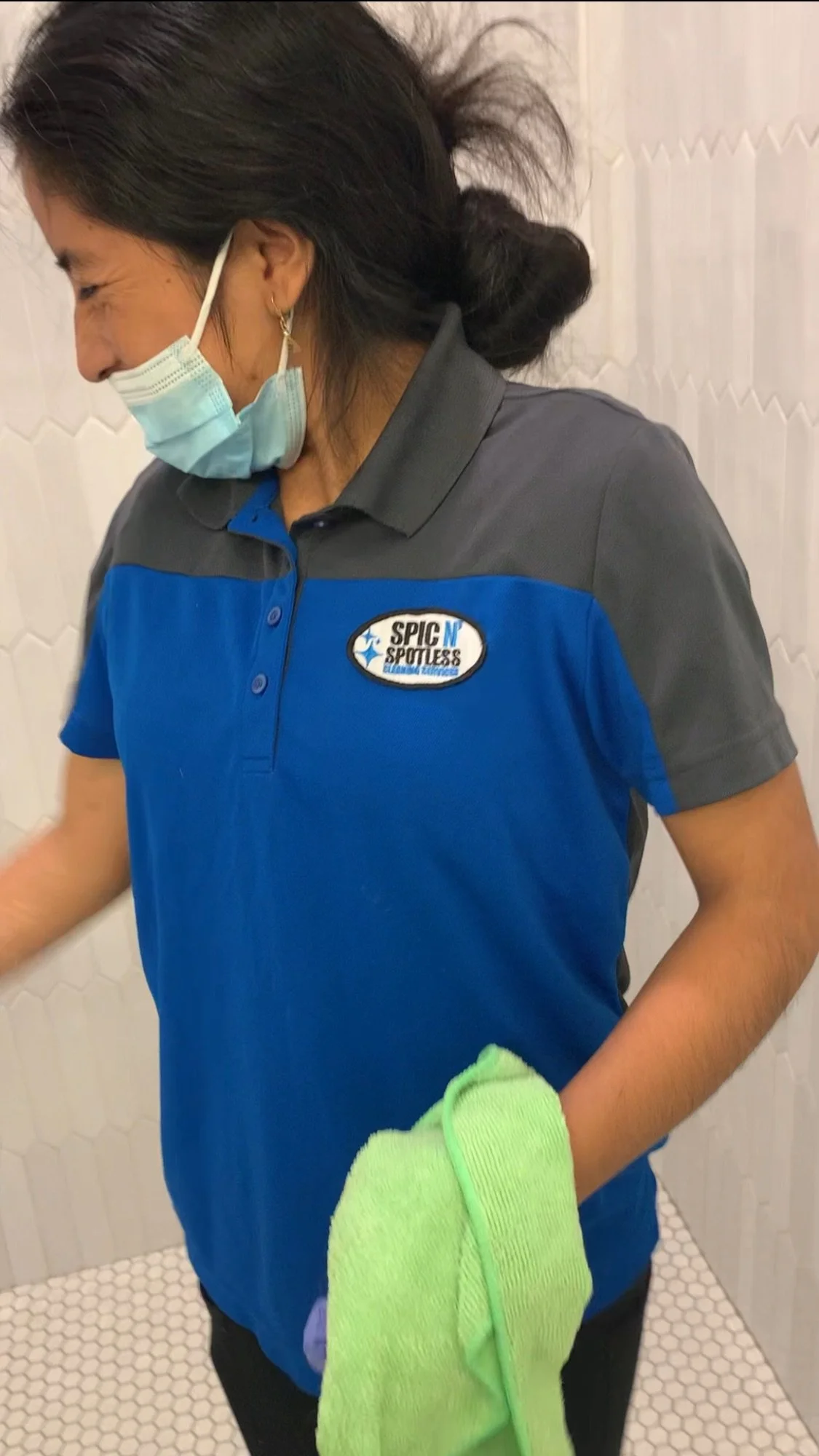 A woman wearing a blue and gray uniform with a logo that reads 'SPIC N SPOTLESS Car Wash' is smiling and holding a green cloth in her right hand. She is also wearing a face mask and standing in a tiled room.