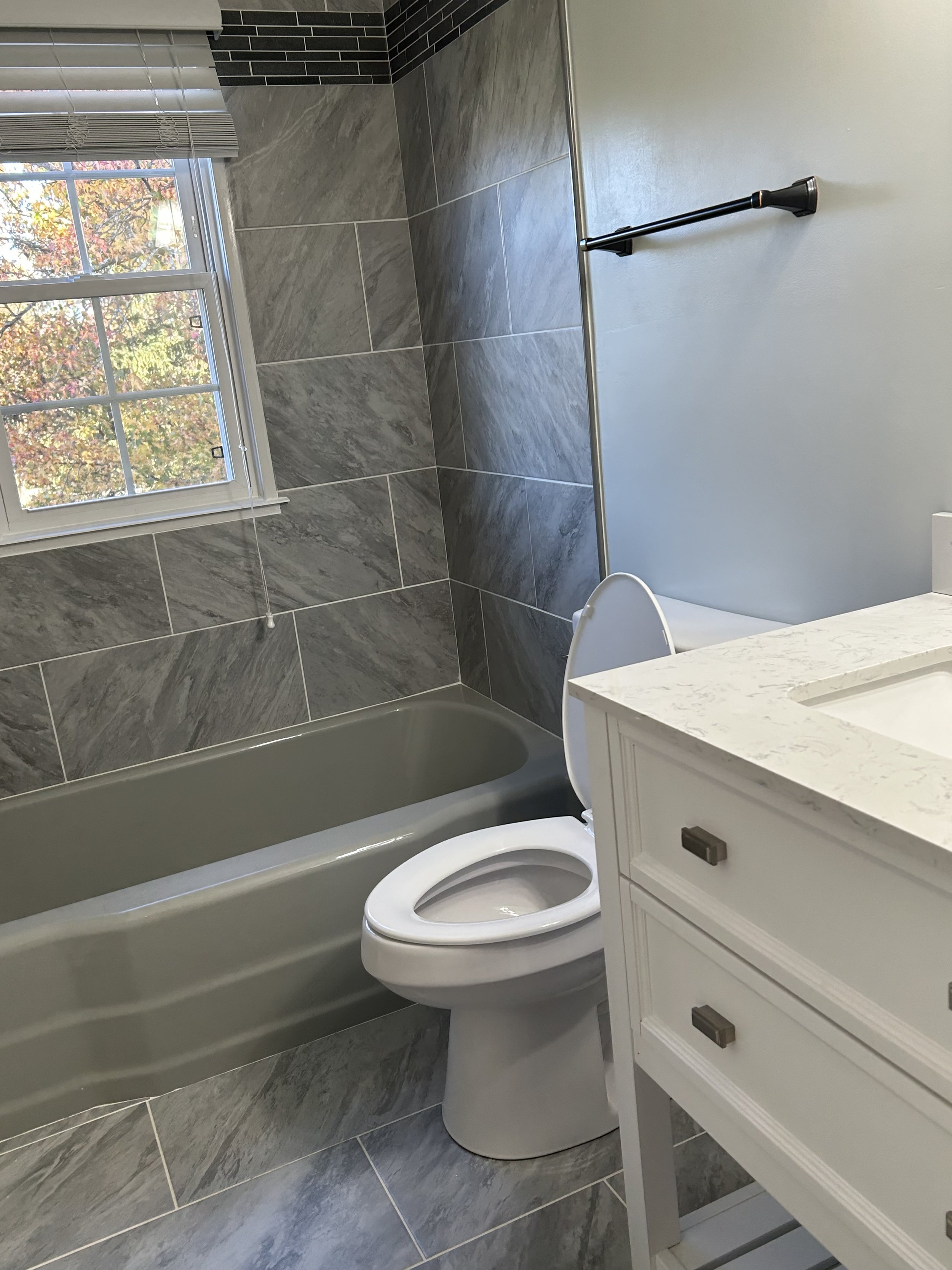 Bathroom with gray tiled walls and floor, white vanity cabinet with marble countertop, toilet, bathtub, window with blinds, and towel bar.