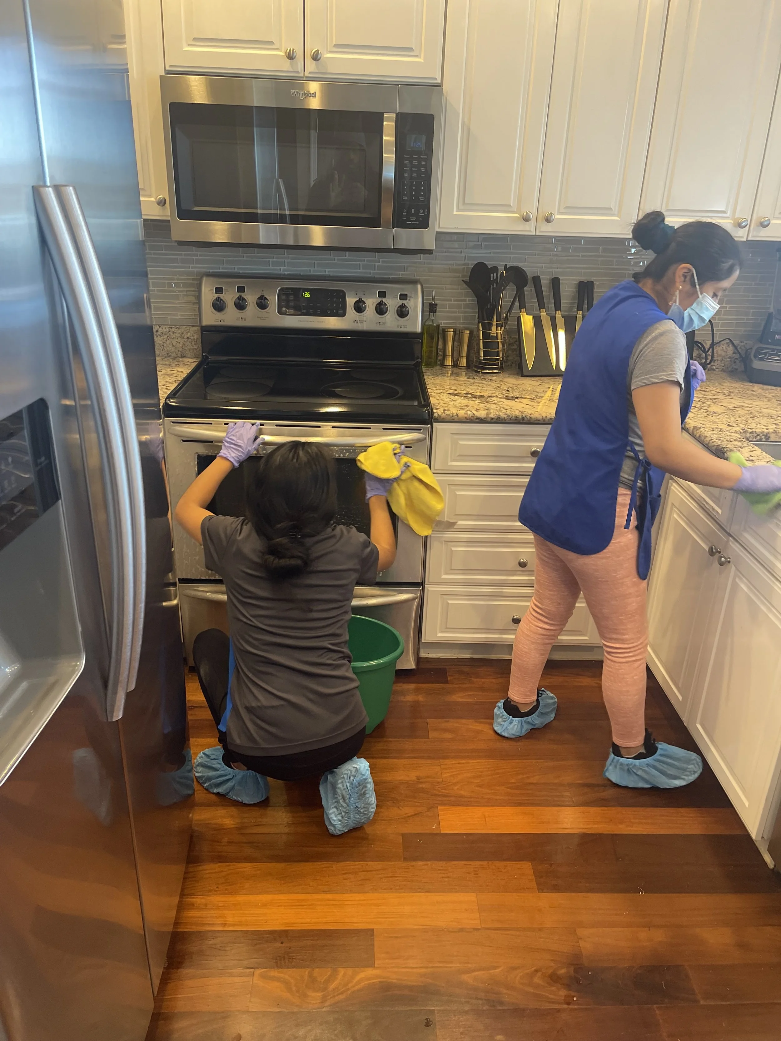 A woman and a child are cleaning a kitchen. The woman is standing by the counter, wearing a mask, blue apron, and shoe covers. The child is crouched near the open oven, wearing shoe covers and gloves, holding a yellow cleaning cloth, and facing away from the camera.