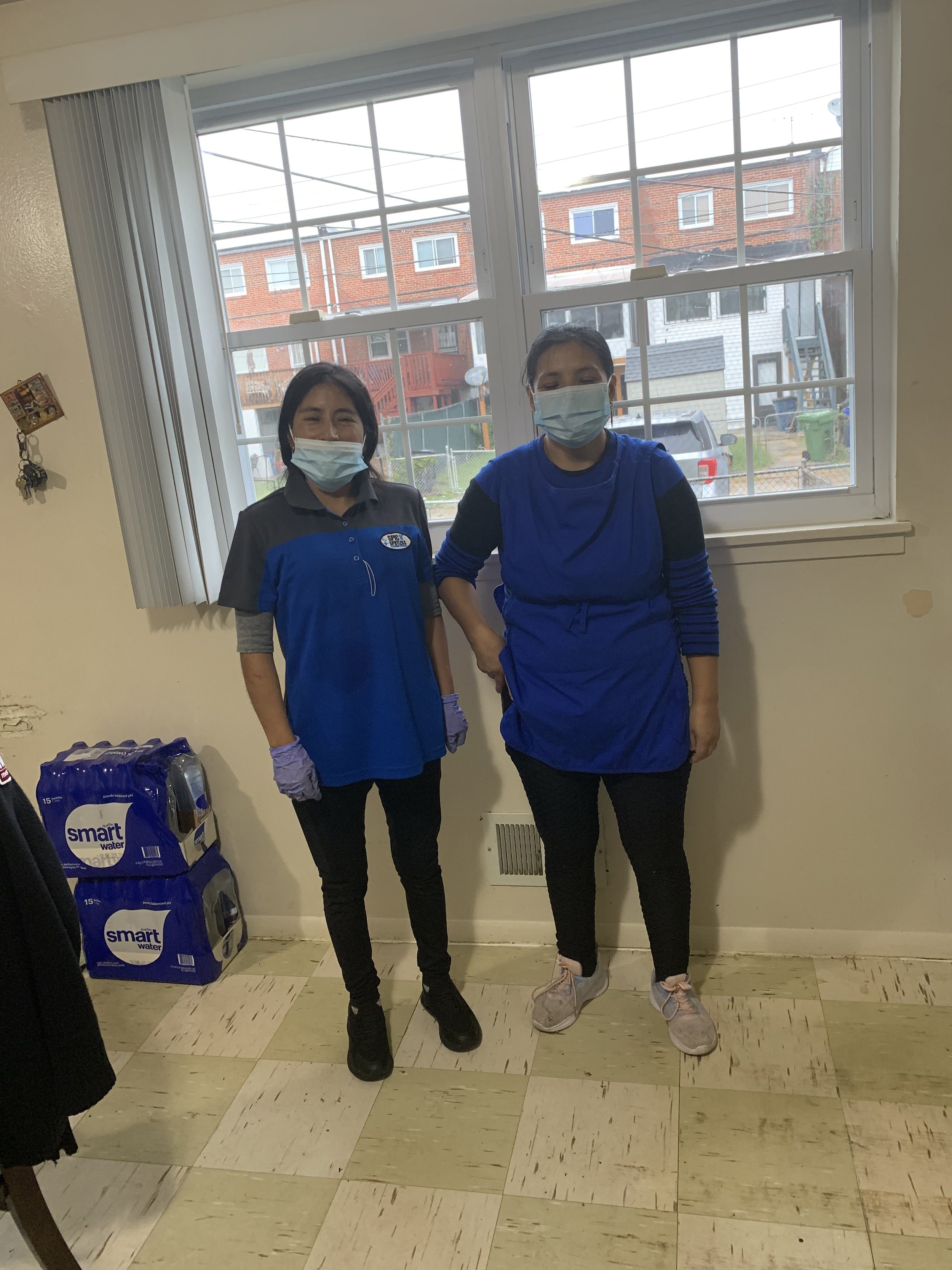 Two women wearing face masks standing indoors in front of a large window, with houses and a car visible outside. One woman wears a gray and blue uniform, and the other wears a blue top and black pants.