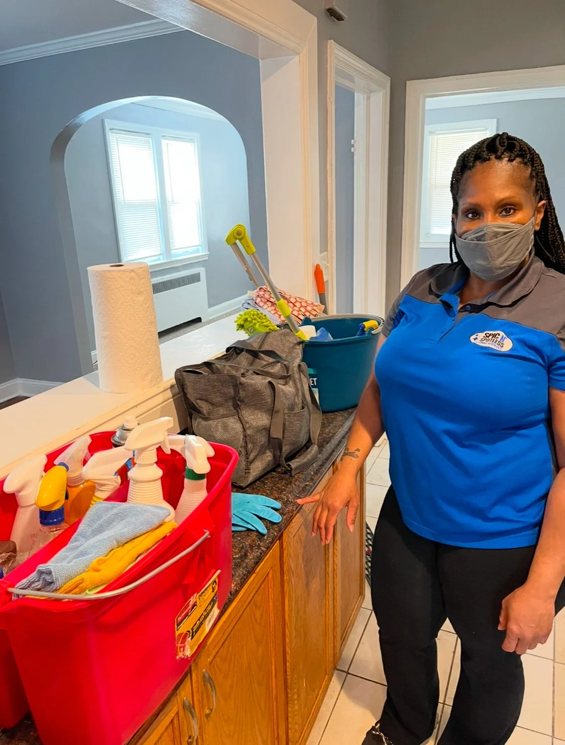 A woman wearing a gray face mask and a blue uniform stands in a kitchen next to cleaning supplies and equipment, including spray bottles, gloves, and a bucket.