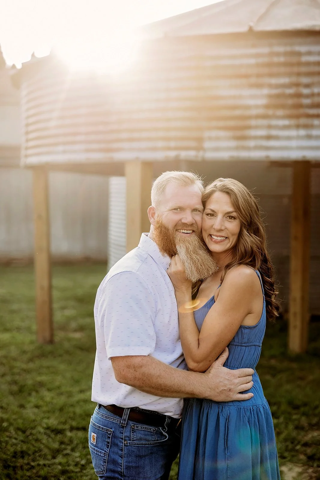 A couple embracing and smiling outdoors with a rustic metal structure in the background, under warm sunlight.