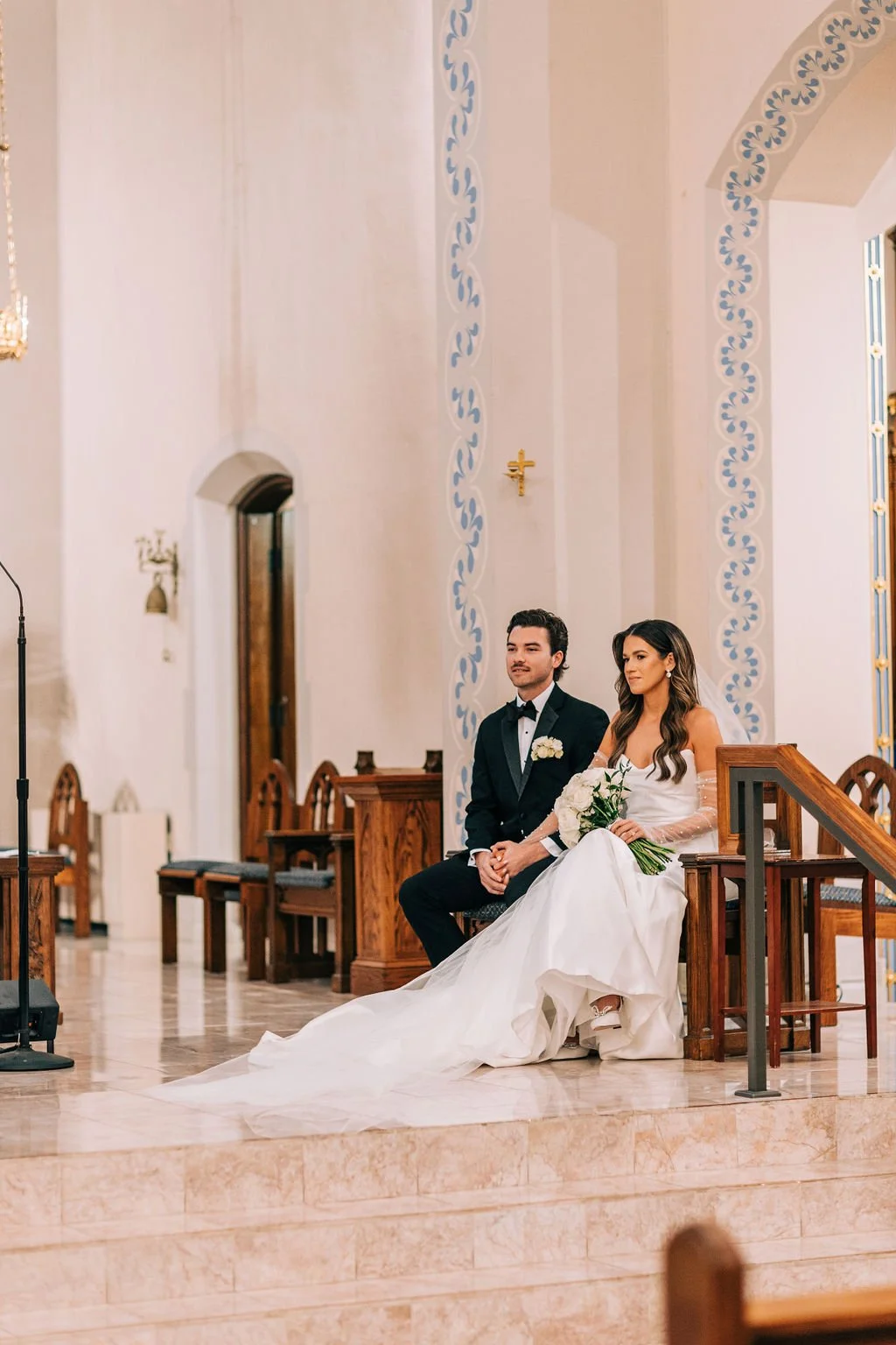 Bride and groom sitting in a church holding hands during a wedding ceremony, with elegant decorations and attire.