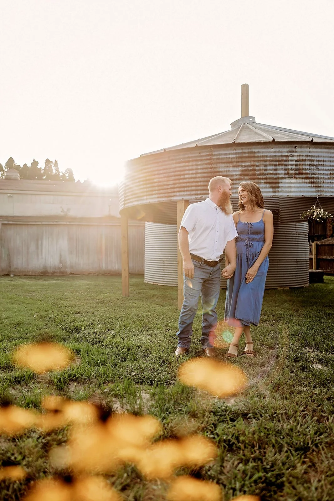 A couple standing and holding hands in a grassy field near a metal grain silo, backlit by the setting sun. The woman is in a blue dress, the man in a white shirt and jeans. Foreground shows blurred yellow flowers.
