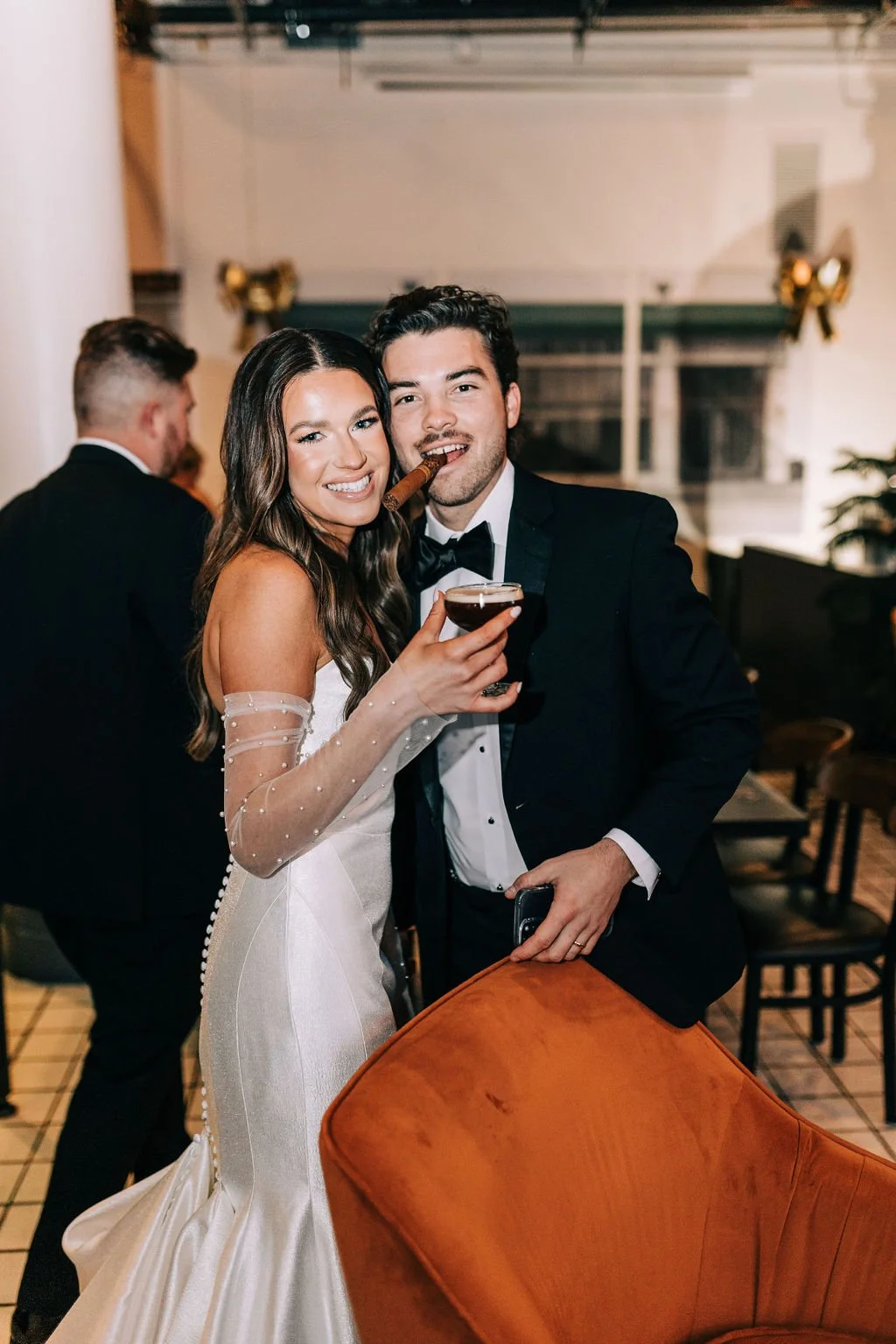A couple in formal attire smiling, the woman in a white gown and the man in a tuxedo with a cigar and a drink, at an indoor event.