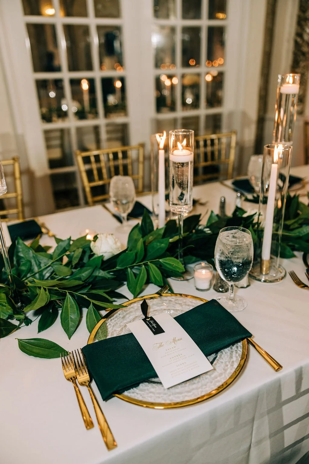 Elegant table setting with gold cutlery, green napkins, and menu card on a glass plate. Greenery decorates the table alongside tall candles and glass goblets. Gold chairs and windowed background visible.