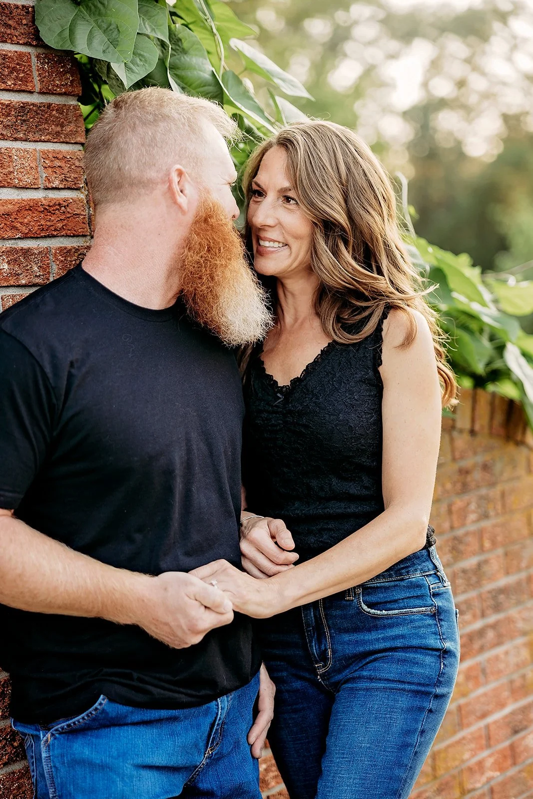 A man and woman standing closely together, smiling at each other, near a brick wall with green leaves overhead. The man has a long red beard and is wearing a black t-shirt, while the woman is wearing a black lace top and jeans.