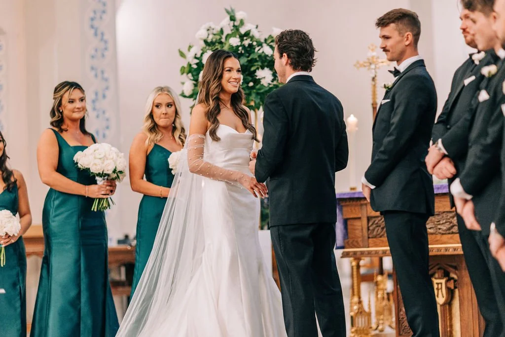 A bride and groom standing at the altar during a wedding ceremony, surrounded by bridesmaids in teal dresses and groomsmen in black suits, in a church setting.