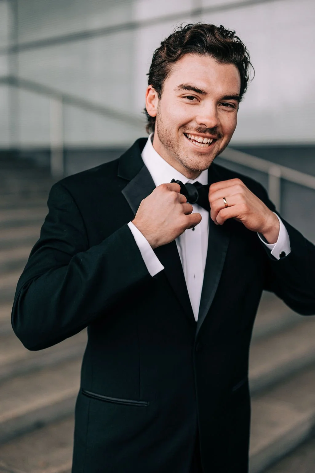 Man in a tuxedo adjusting his bow tie while smiling in a formal setting.