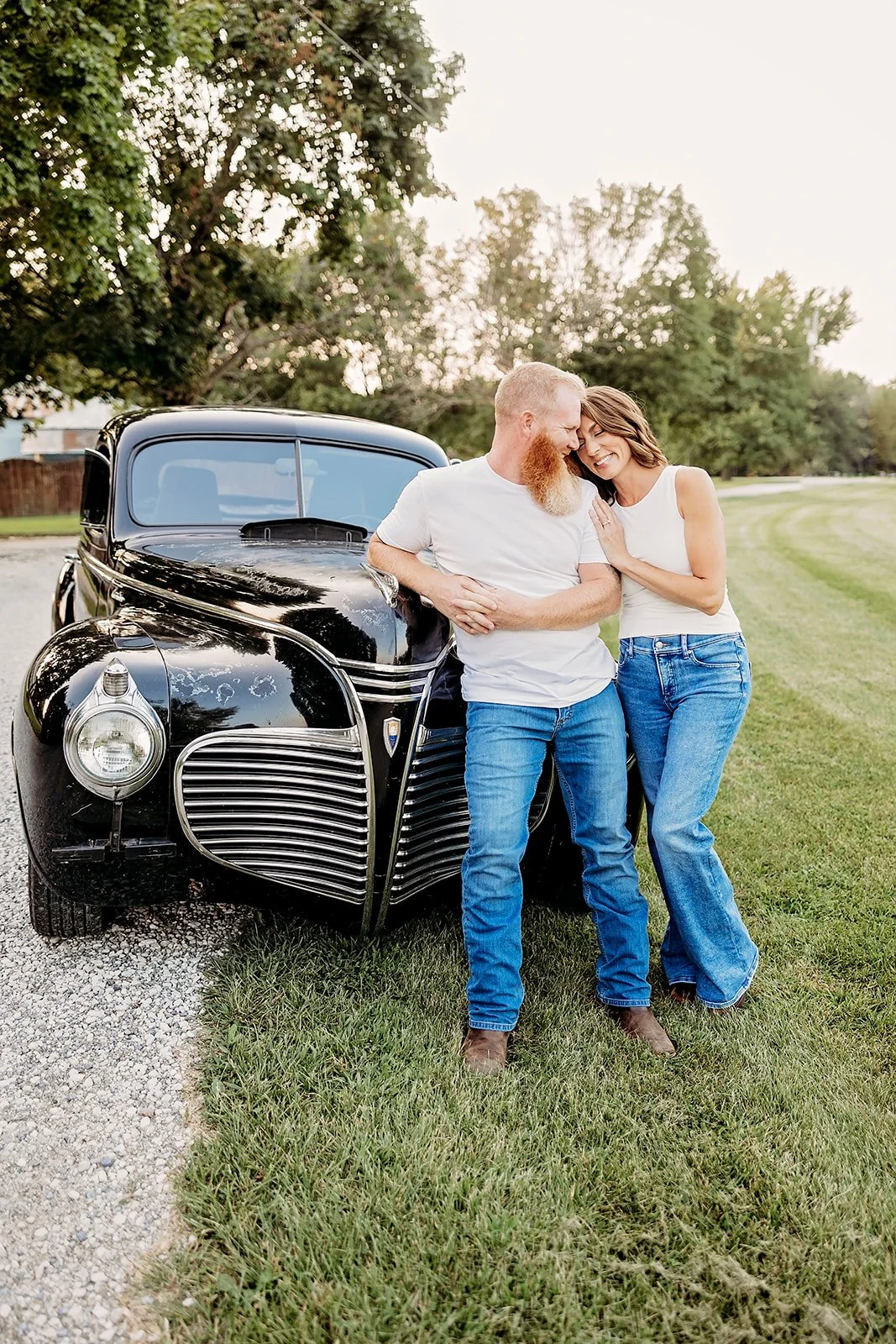 Couple leaning on a vintage black car in a grassy area.