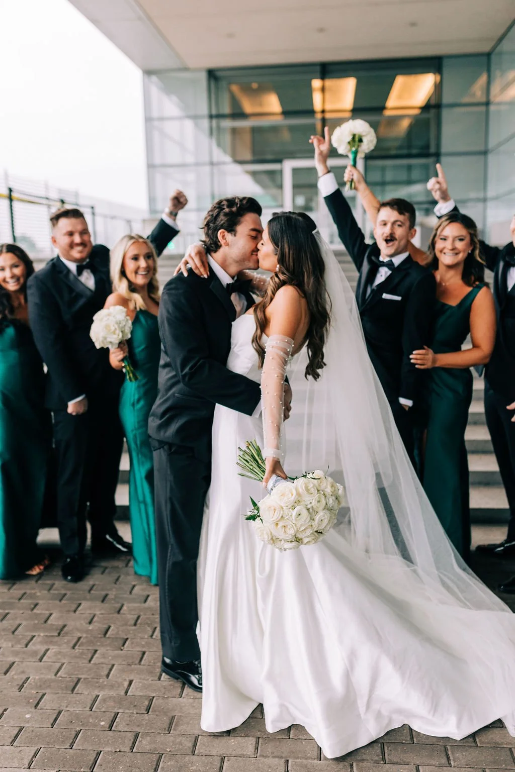 Bride and groom kissing with wedding party cheering, dressed in formal attire and holding bouquets.