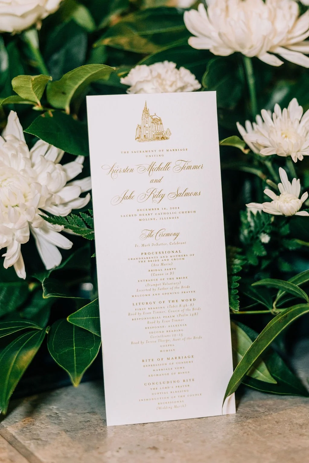 Wedding ceremony program surrounded by white flowers and green leaves.