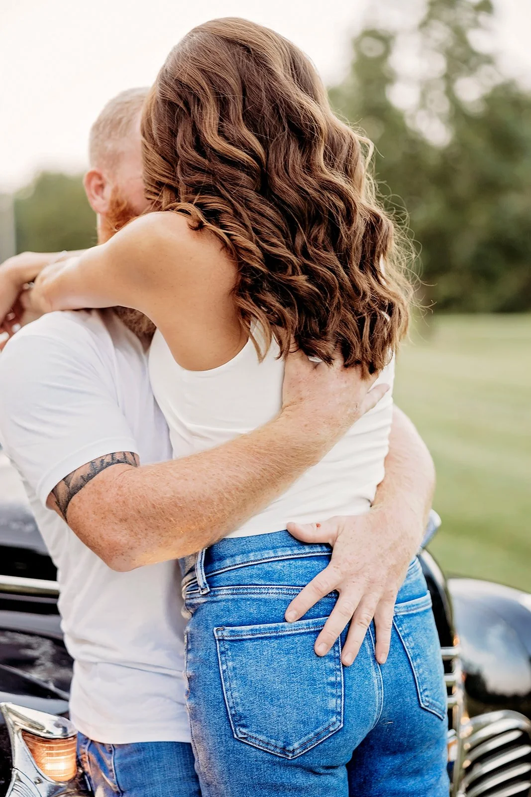 Couple embracing near a vintage car in a grassy area, woman with long wavy hair and wearing jeans, man in white T-shirt with tattooed arm.