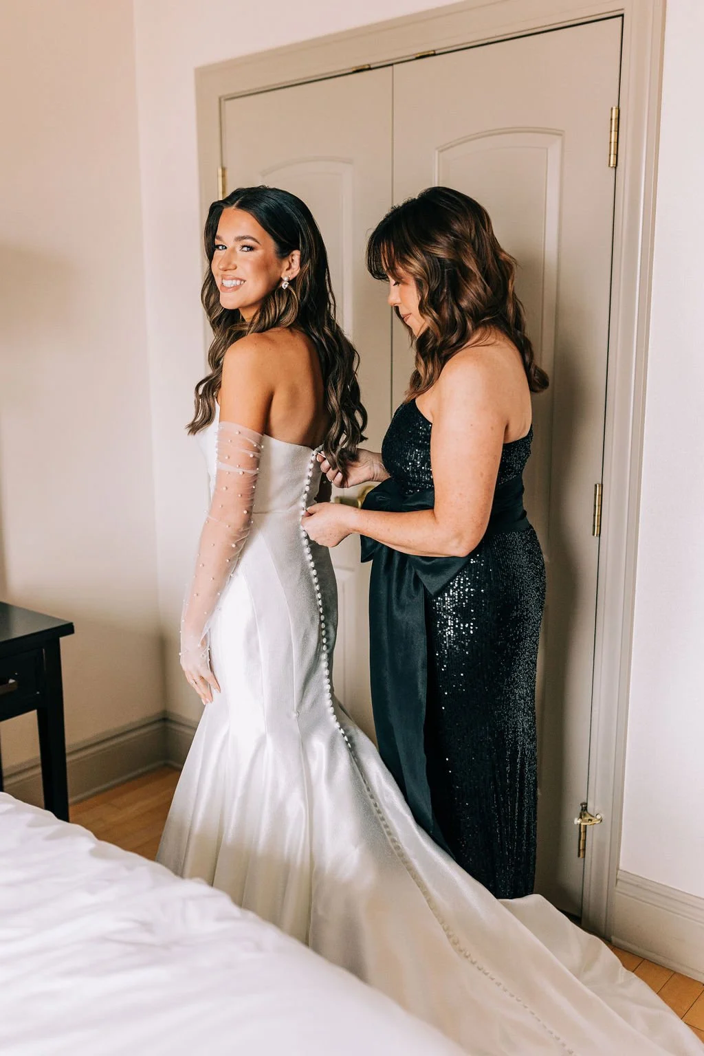 A bride in a white wedding dress being assisted by another woman wearing a black dress. The bride is smiling in a room with closed double doors and a bed visible.