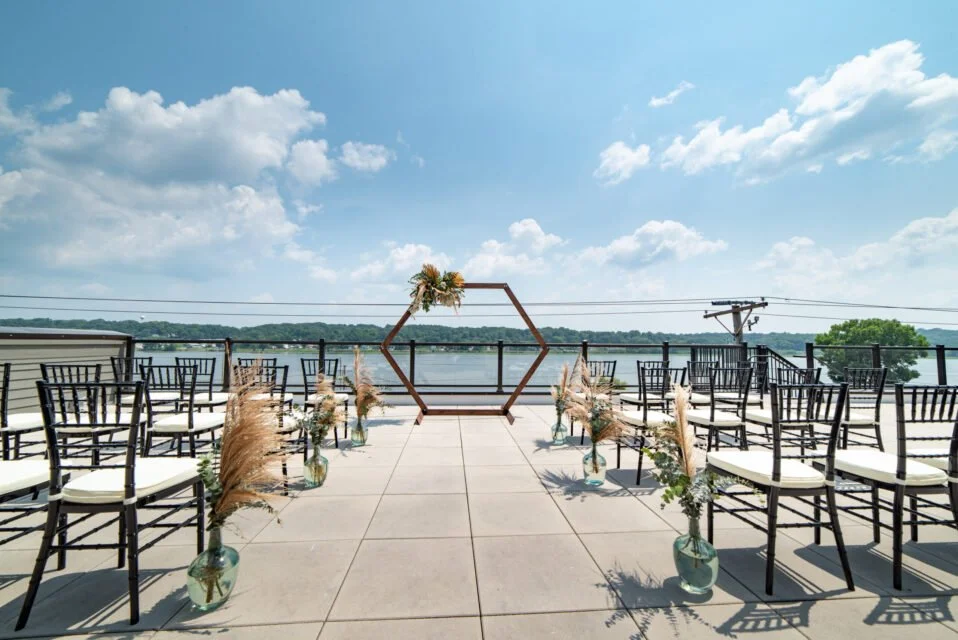 Outdoor wedding ceremony setup with hexagonal arch and black chairs overlooking a lake.