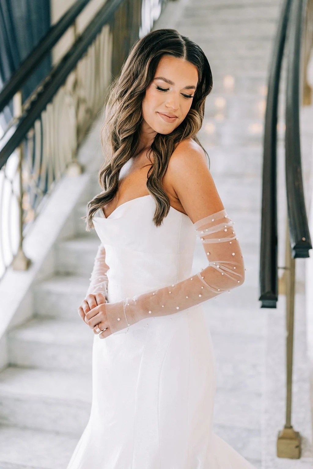 Bride in a white wedding dress with sheer, pearl-embellished sleeves, standing on a marble staircase.