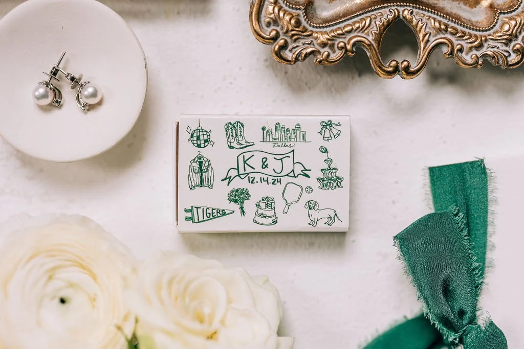 Flat lay of wedding invitation suite with personalized box, pearl earrings, ornate frame, green ribbon, and white roses on a light surface.