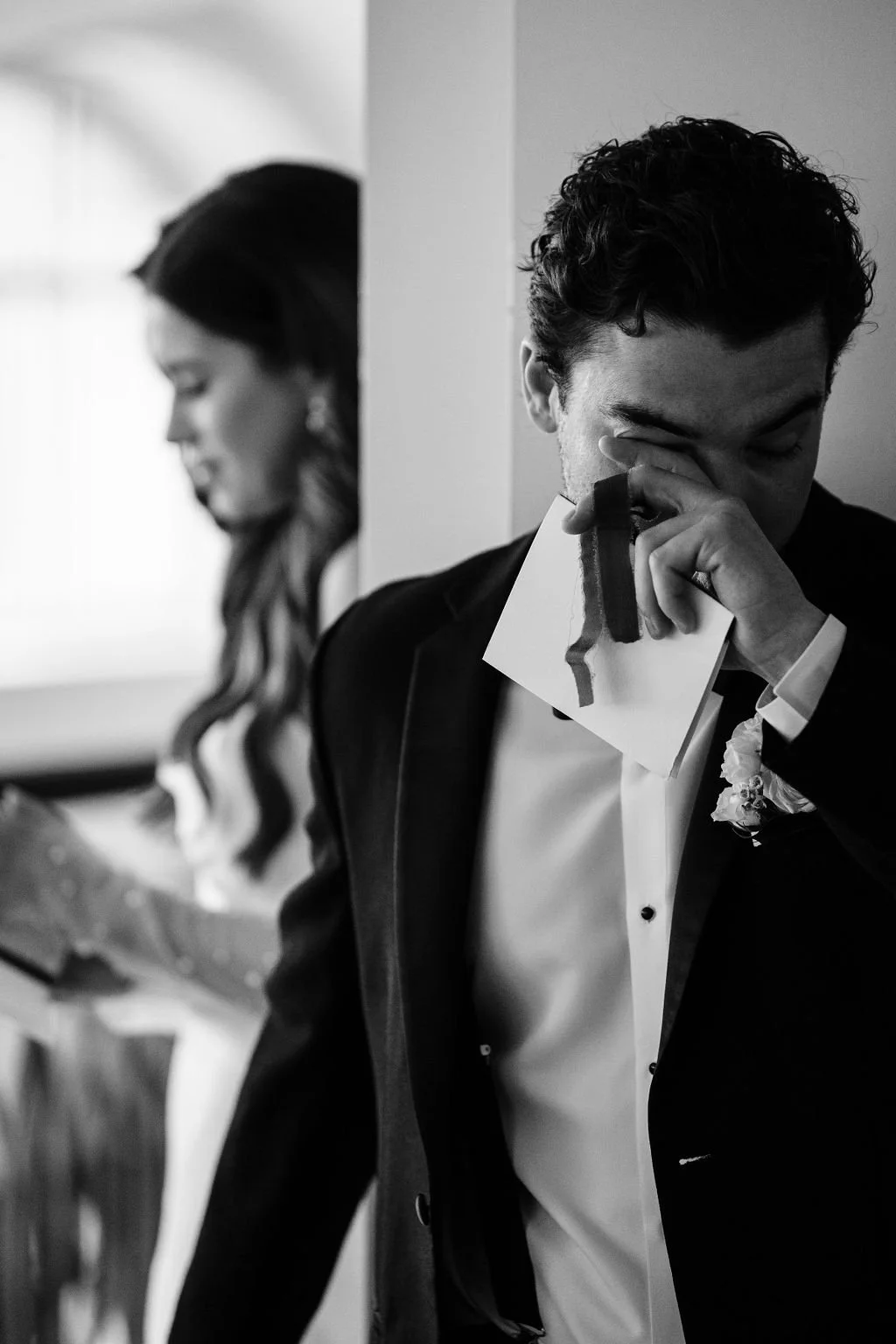 A bride and groom stand back to back on either side of a doorway, each holding a letter. The groom wipes his eyes with one hand, appearing emotional, while the bride looks down at her letter. The scene is captured in black and white, highlighting an