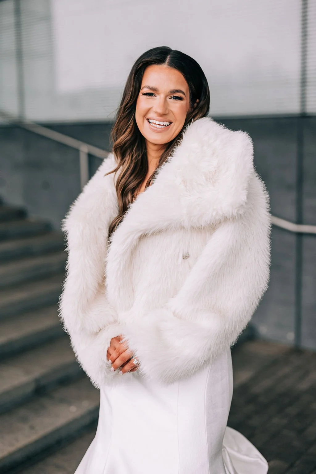 Woman in white fur coat standing on stairs, smiling.