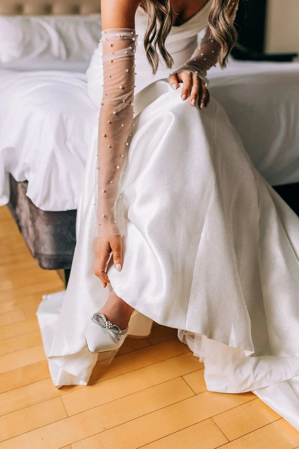 Bride in white dress with pearl-accented sheer sleeves adjusting jeweled shoe.